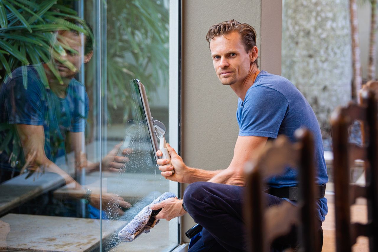 A man wearing a blue t-shirt cleaning a large glass pane indoors with a squeegee.